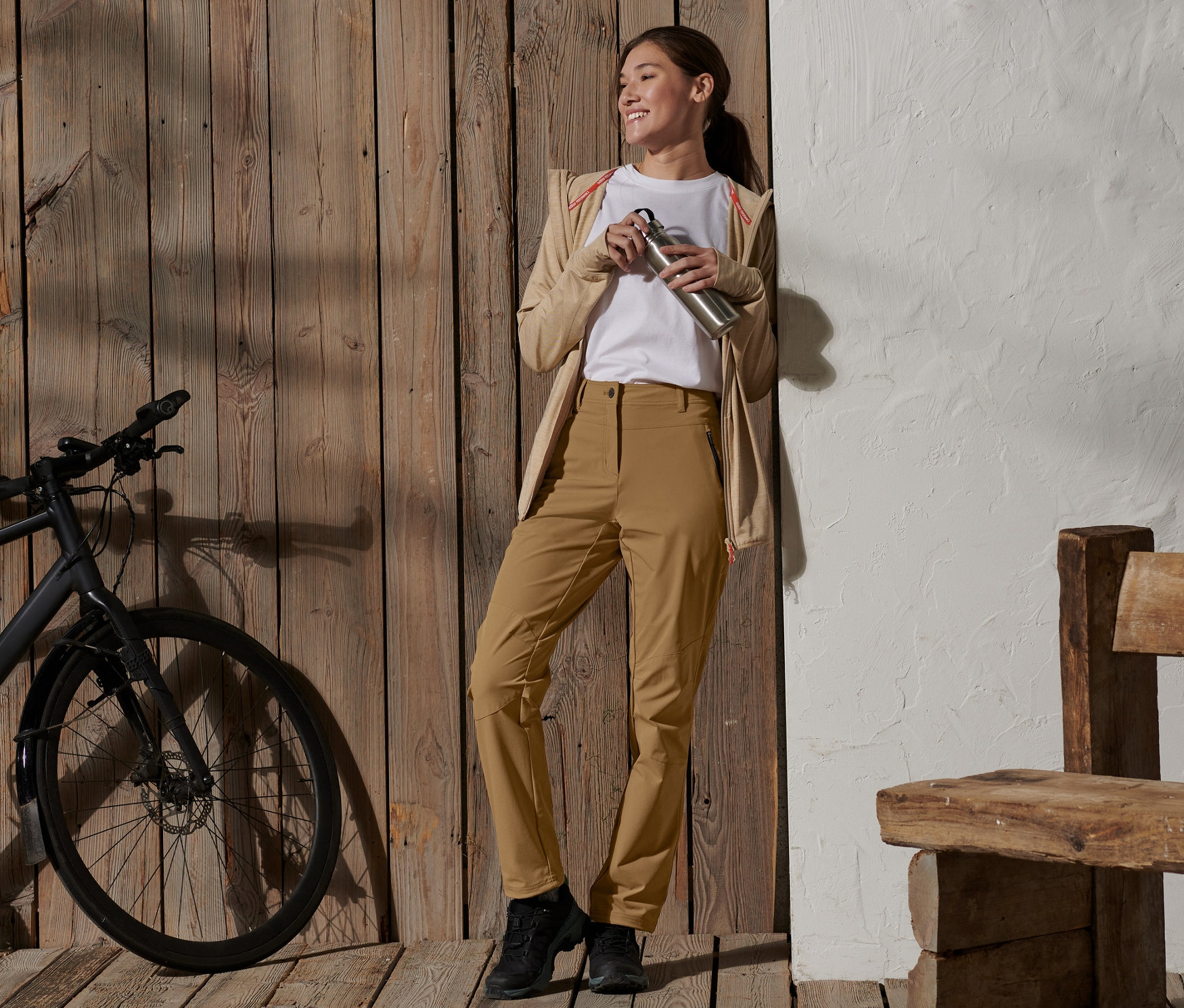 Žena se usmívá a drží nerezovou láhev na vodu, opírá se o dřevěnou stěnu a má na sobě softshellové kalhoty.