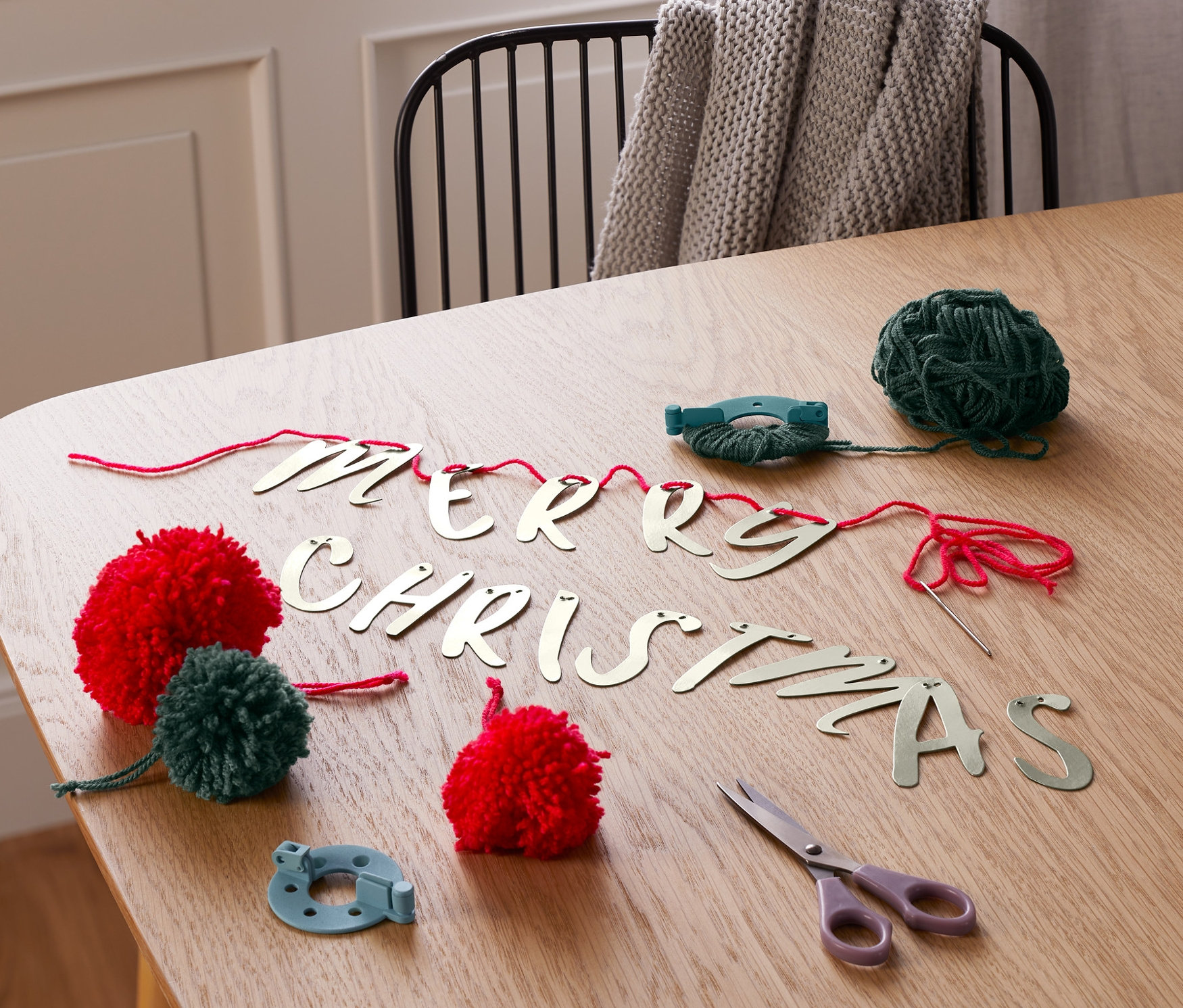 Na stole leží sada na výrobu girlandy s bambulkami. Jsou zde červené a zelené bambulky, zelená příze, nůžky a navlečená slova "MERRY CHRISTMAS".