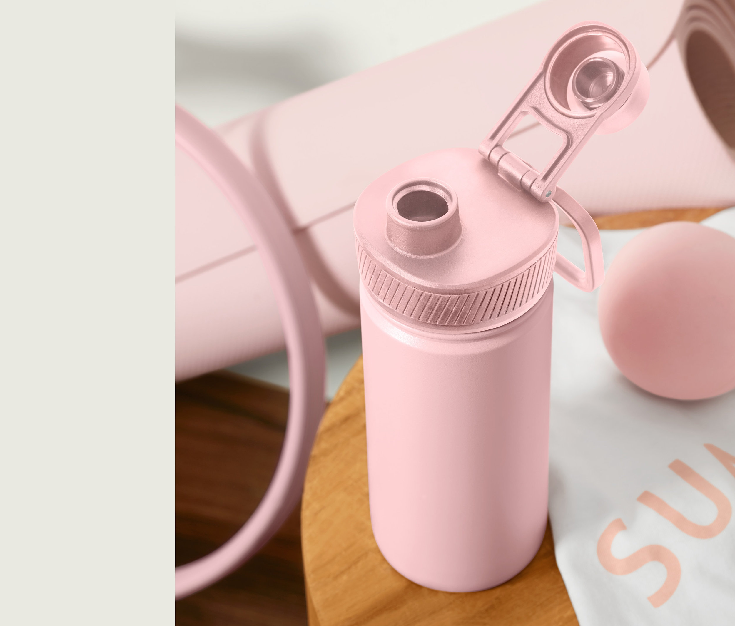 Růžová sportovní láhev na pití s otevřeným víčkem stojí na dřevěném stole vedle srolované podložky na cvičení a masážního míčku.