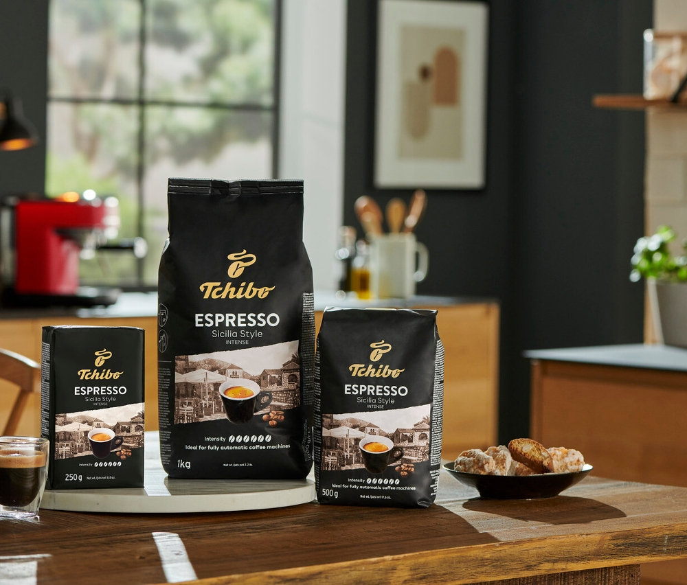 Tři balení kávy Tchibo Espresso Sicilia Style (1 kg, 500 g, 250 g) stojí na dřevěném stole vedle šálku espressa a talíře sušenek.
