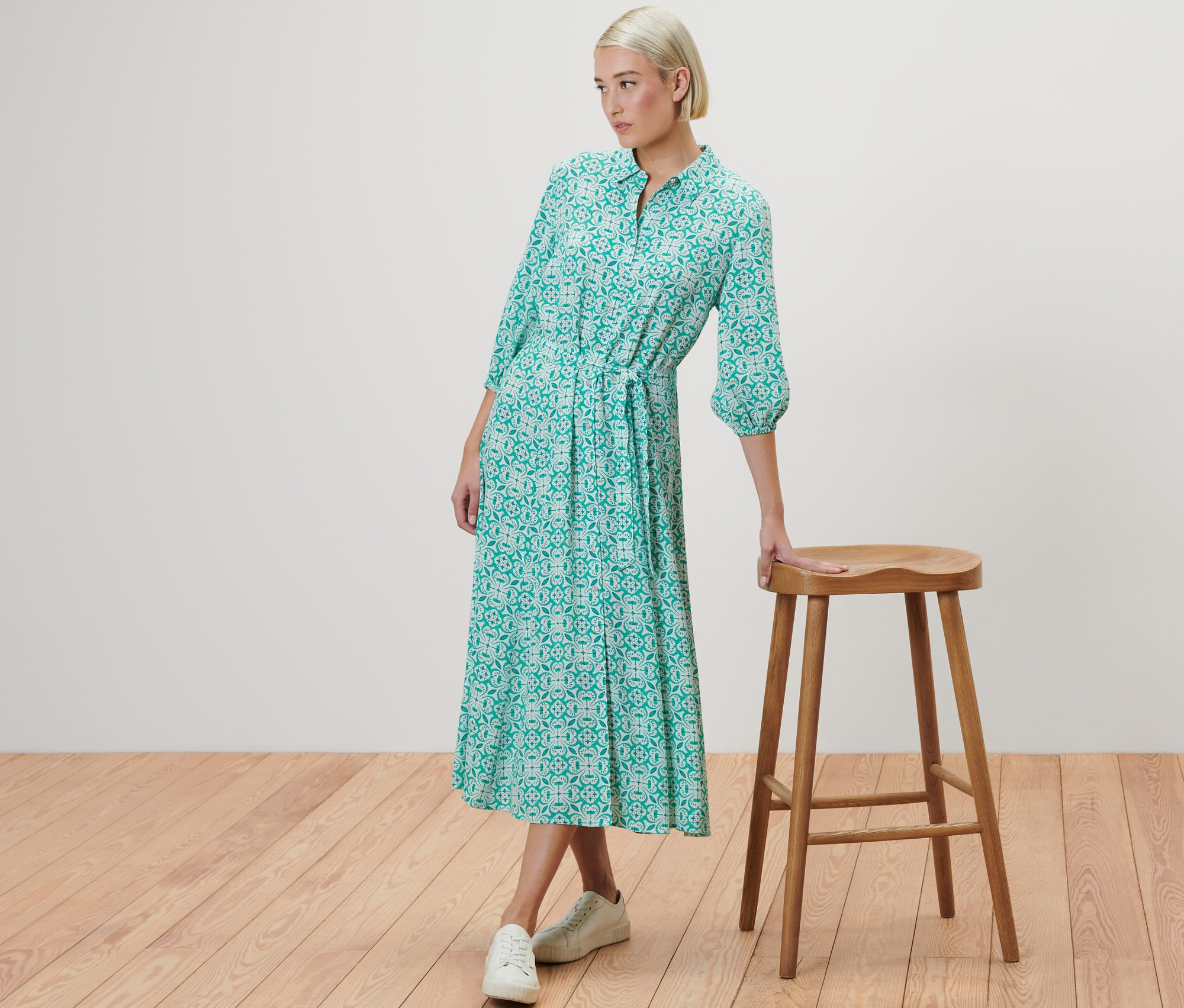 Žena pózuje vedle dřevěné stoličky a má na sobě halenkové šaty s celoplošným potiskem.