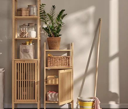 V místnosti stojí dva úložné regály ve skandinávském designu, naplněné úložnými sklenicemi, košíky a textilem. Je zde také koš na prádlo a vědro s mopem.
