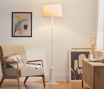 Bílá stojací lampa s textilním stínidlem stojí v obývacím pokoji vedle béžového křesla.