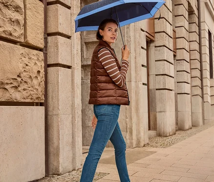 Žena kráčí po chodníku s deštníkem, má na sobě oboustrannou prošívanou vestu, džíny slim – Fit »Emma«, tmavě modré a kožené tenisky.