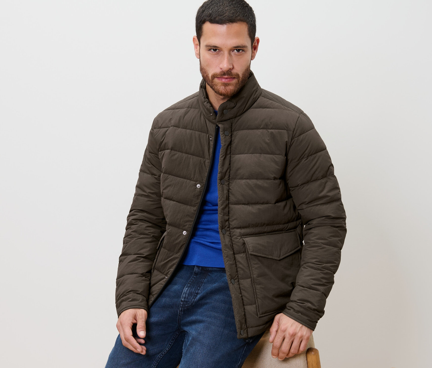 Muž sedí a má na sobě prošívanou bundu, modré džíny a modré tričko.