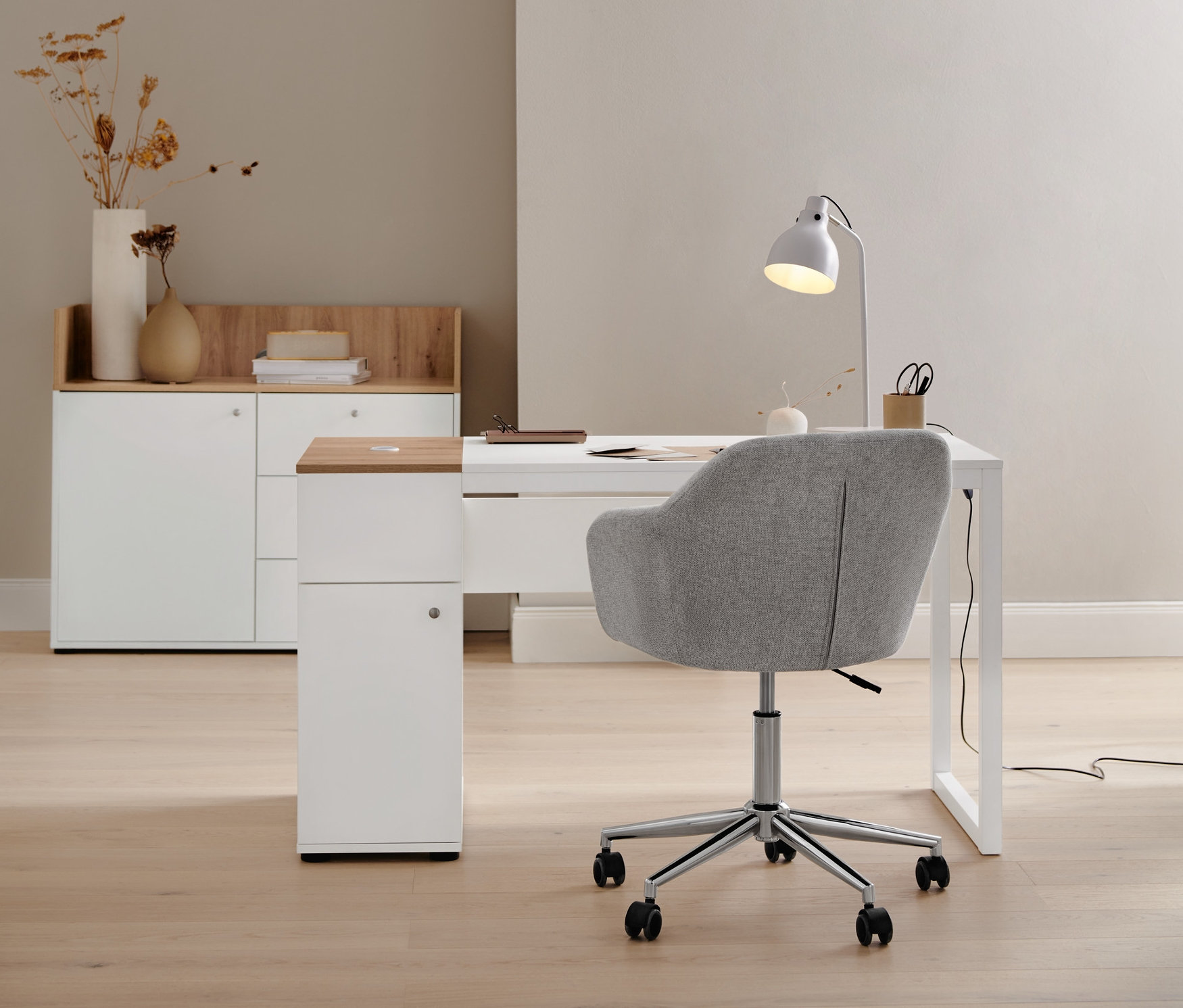 Béžová kancelářská židle stojí u psacího stolu s úložným prostorem. V pozadí je komoda se zásuvkami a dvířky a kovová stolní lampa.