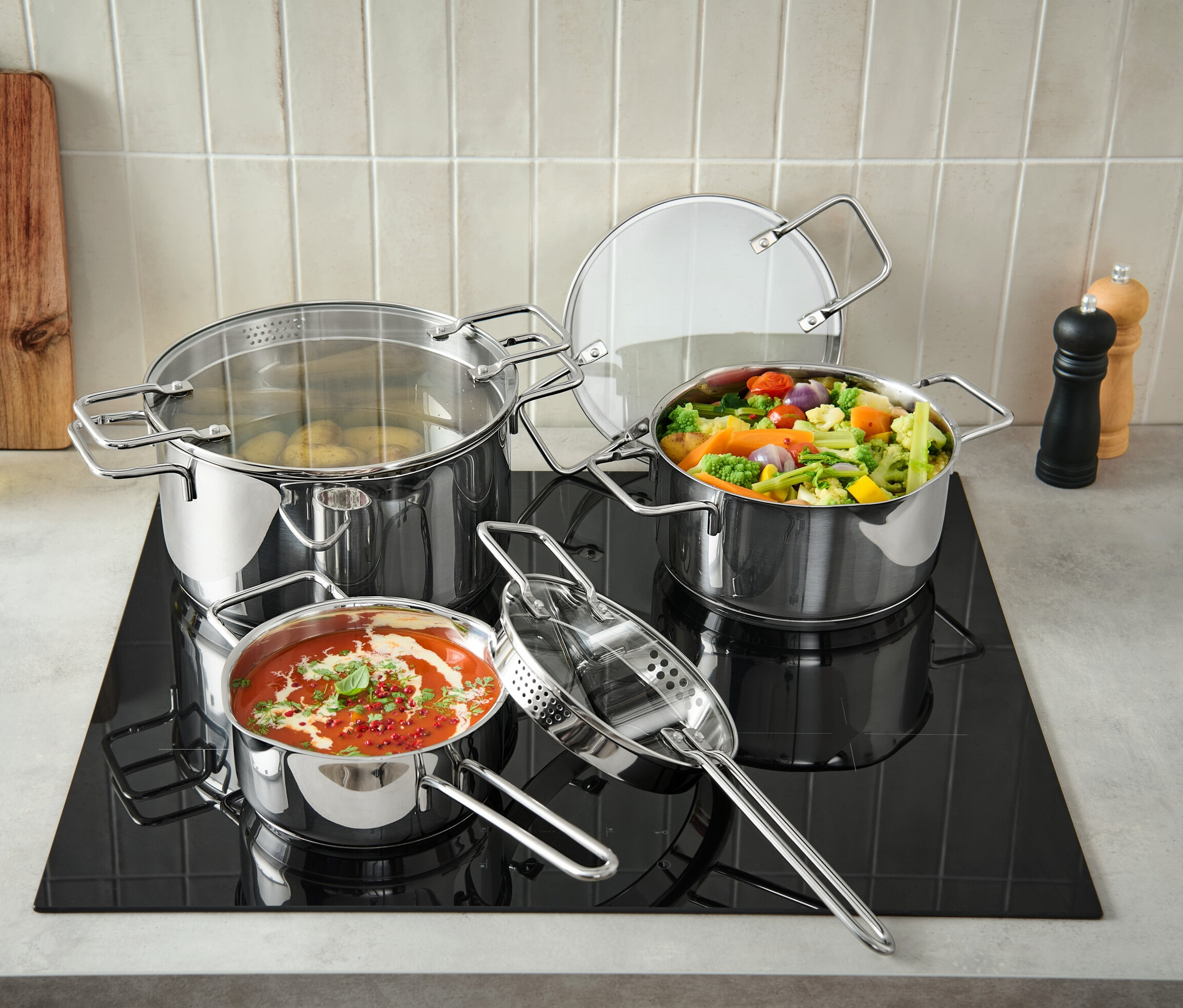 Tři profesionální hrnce z ušlechtilé oceli stojí na černé varné desce, jeden hrnec je plný brambor, druhý zeleniny a třetí rajské omáčky.