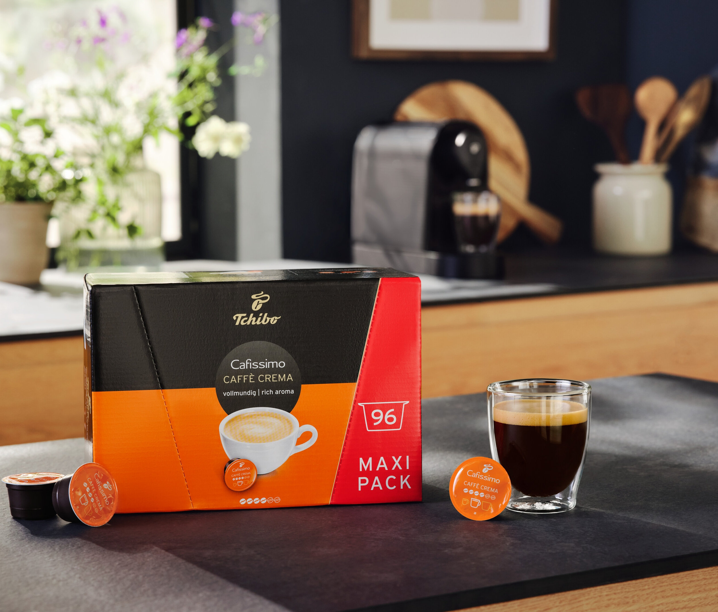 Balení Cafissimo Caffè Crema vollmundig 96 kapslí stojí vedle sklenice s kávou. V pozadí je kávovar Cafissimo pure+ black.