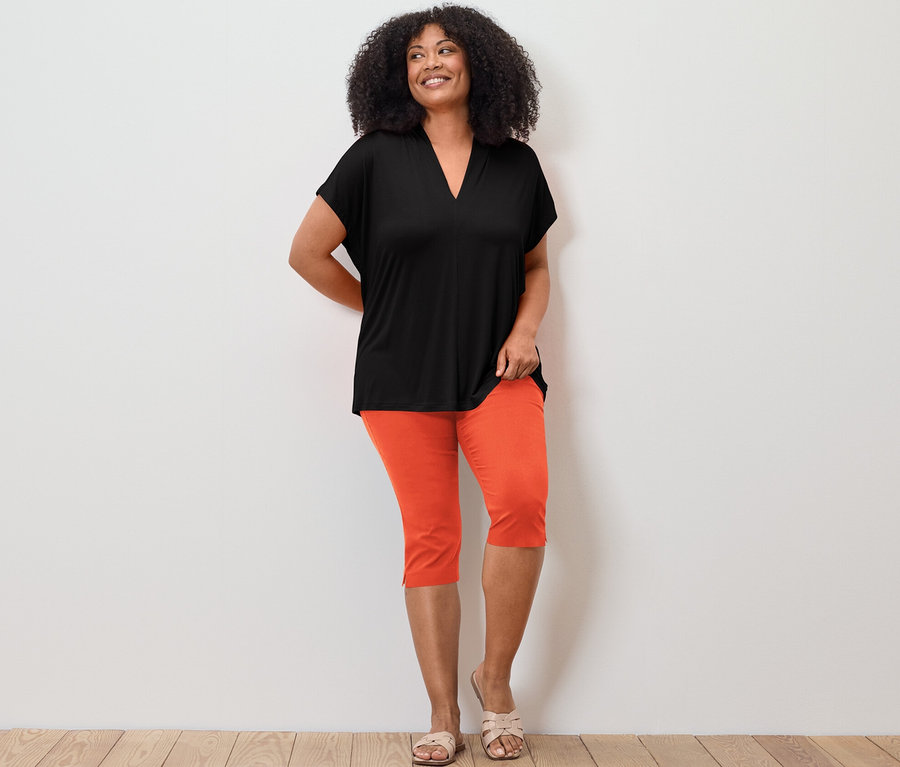 Žena pózuje v černém halenkovém triku s výstřihem do V a oranžových 3/4 strečových kalhotách.