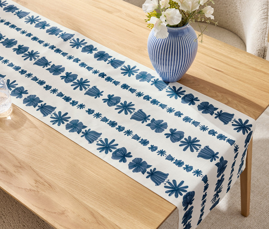 Béžový dřevěný stůl s modrobílým běhounem, modrou vázu s bílými květinami a sklenicí.