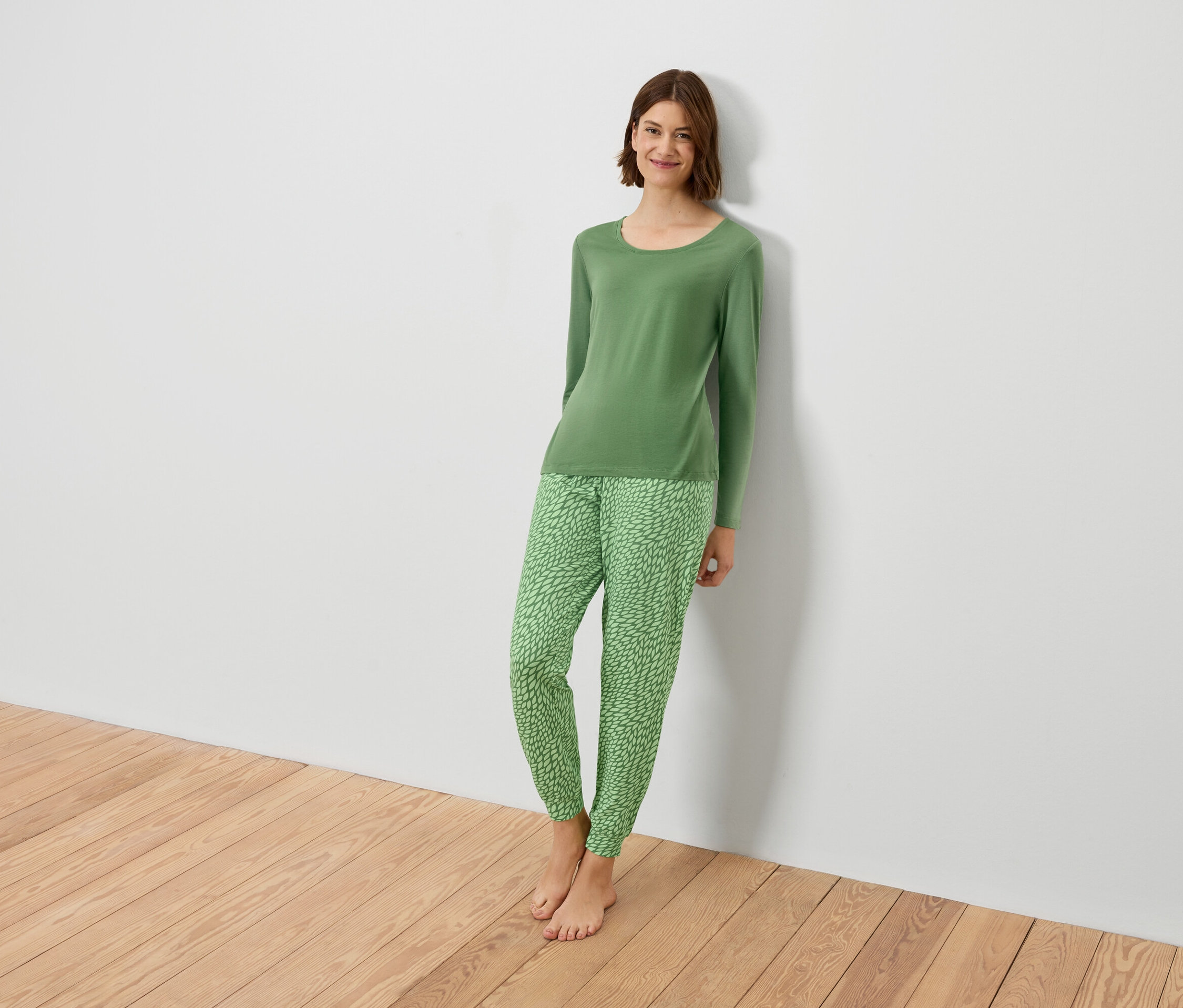Žena v zeleném pyžamu se opírá o bílou zeď.