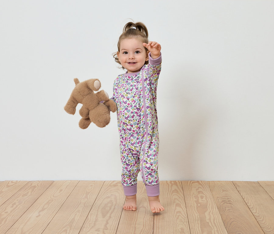 Dítě v květinovém overalu stojí s plyšovým medvídkem.