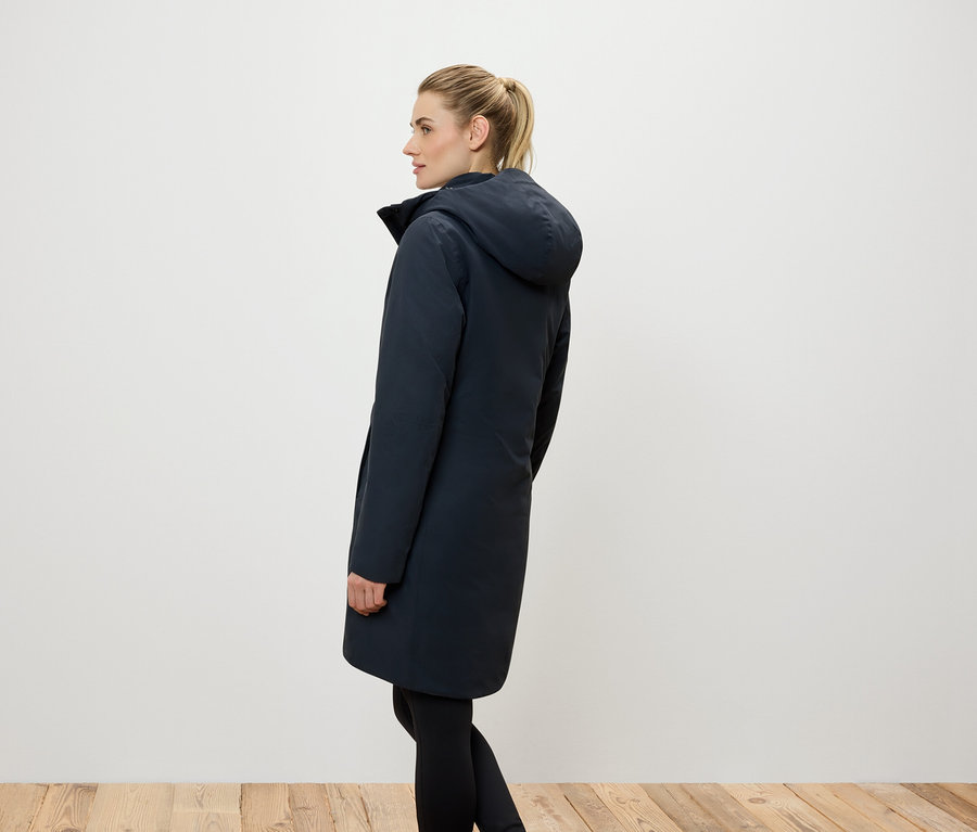 Boční pohled na ženu s blond vlasy staženými do culíku, oblečenou v dlouhém tmavě modrém kabátě s kapucí a černých legínách.