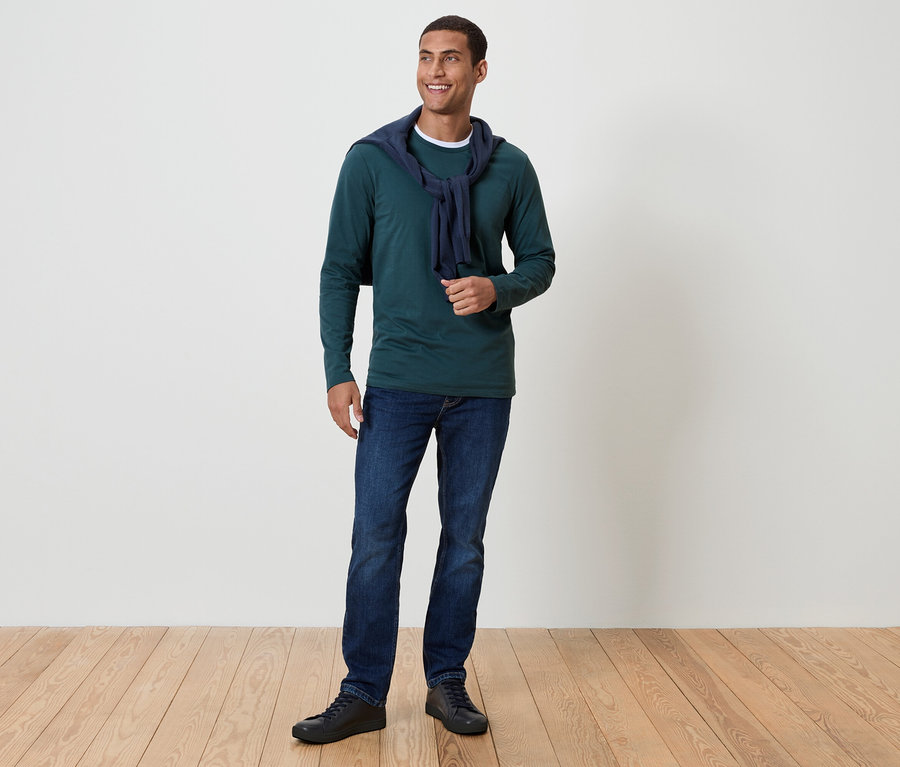 Muž s tmavými vlasy a úsměvem stojí na dřevěné podlaze. Má na sobě zelené tričko s dlouhým rukávem, modré džíny a černé boty. Kolem krku má přehozený tmavě modrý svetr.
