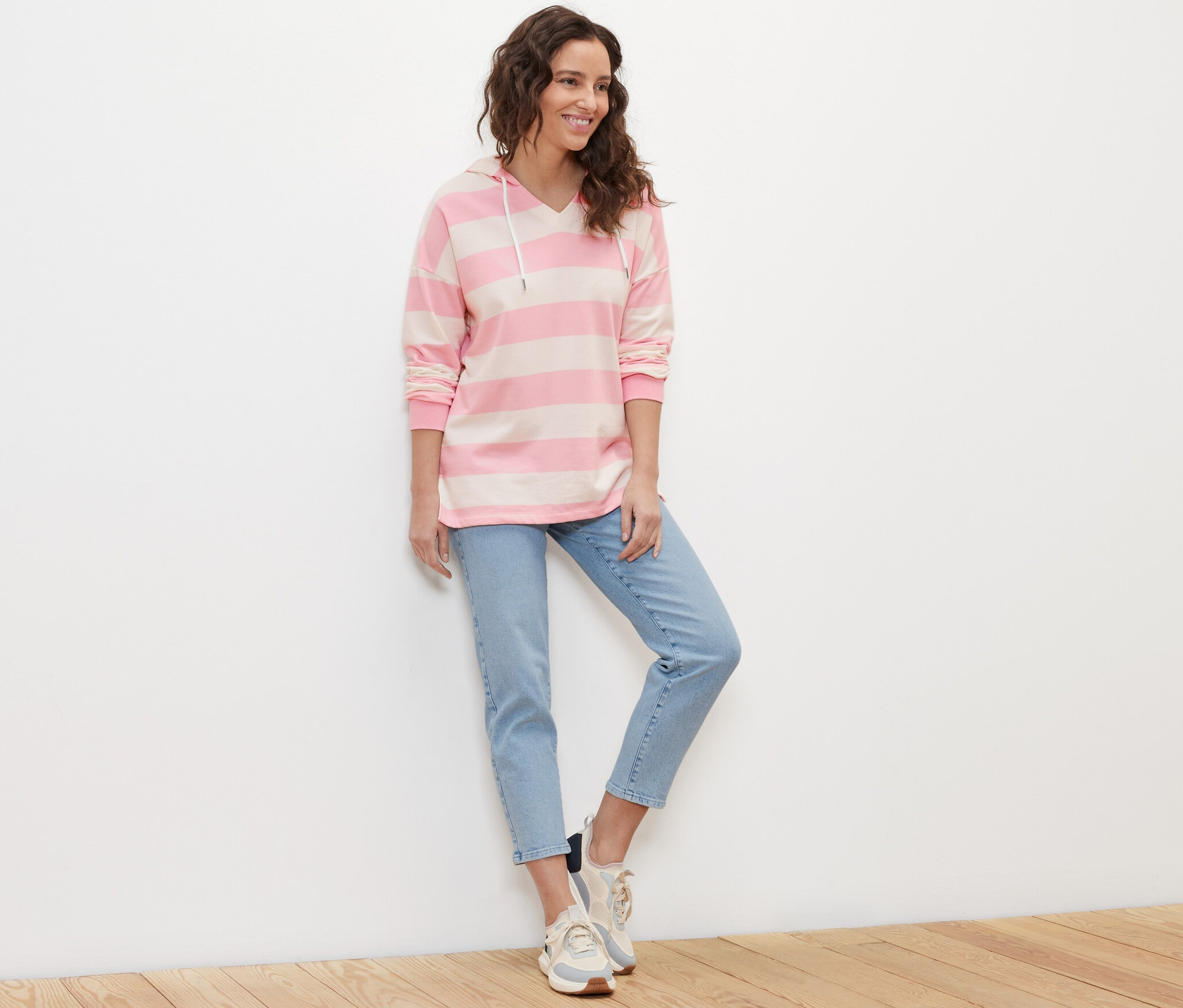 Žena stojí a má na sobě pruhovanou mikinu s kapucí, džíny a tenisky.