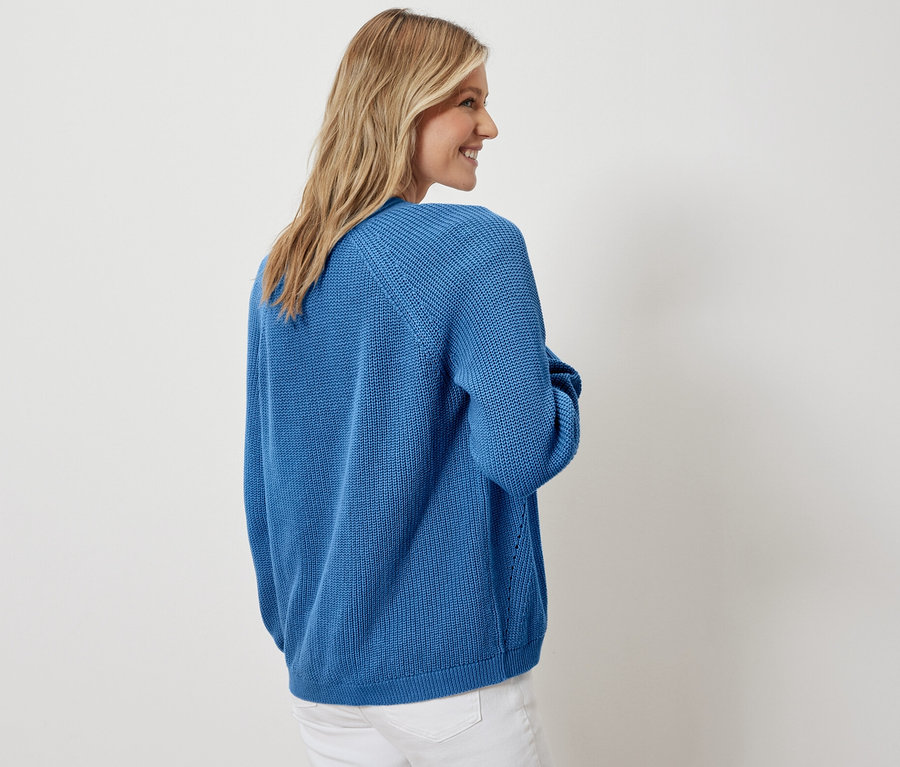 Žena pózuje v otevřeném kardiganu a bílých džínách.