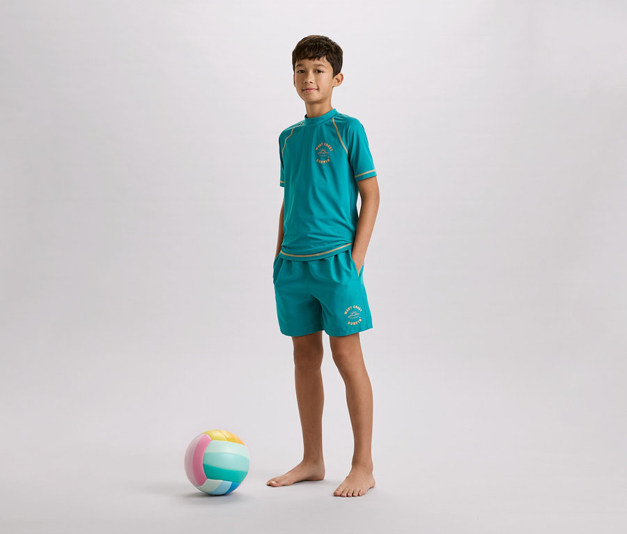 Chlapec stojí bosý v tyrkysovém tričku a kraťasech vedle barevného míče na šedém pozadí.