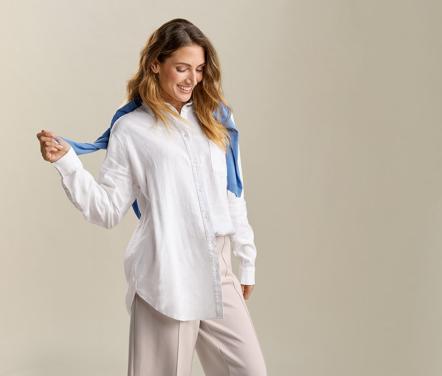 Žena s modrým svetrem přes ramena má na sobě dlouhou bílou lněnou blůzu.