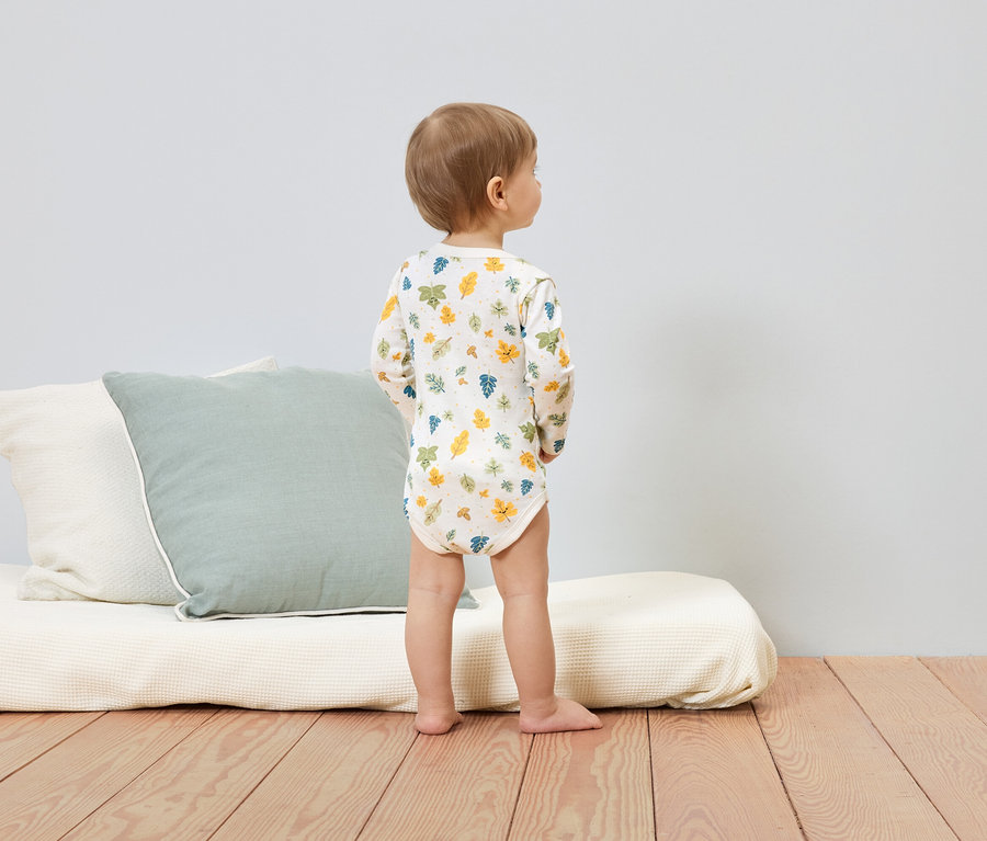 Batole v body s dlouhým rukávem s potiskem listů stojí na dřevěné podlaze vedle polštářů a matrace.