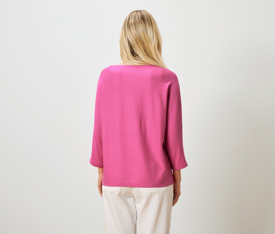 Pohled zezadu na blondýnu v růžovém topu a světlých kalhotách na bílém pozadí.