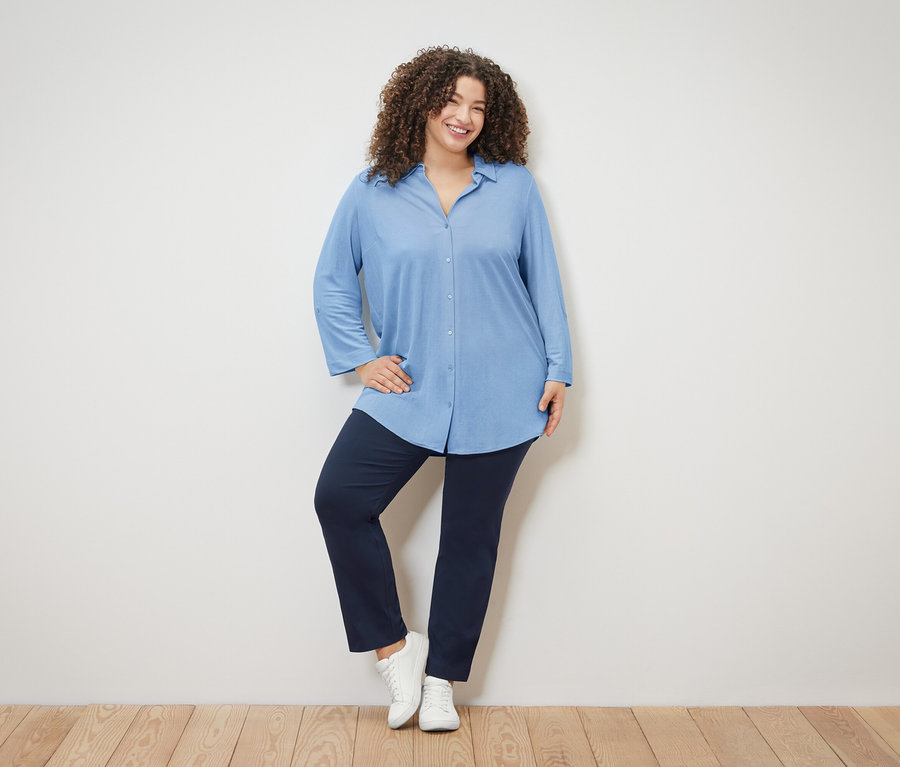 Žena s kudrnatými vlasy má na sobě světle modrou halenku, tmavě modré kalhoty a bílé boty.