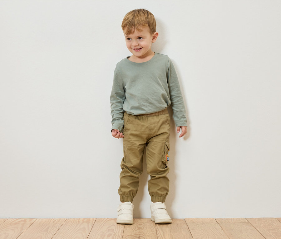 Chlapec s blond vlasy stojí před bílou zdí a má na sobě zelené tričko s dlouhým rukávem, khaki kalhoty a bílé boty.