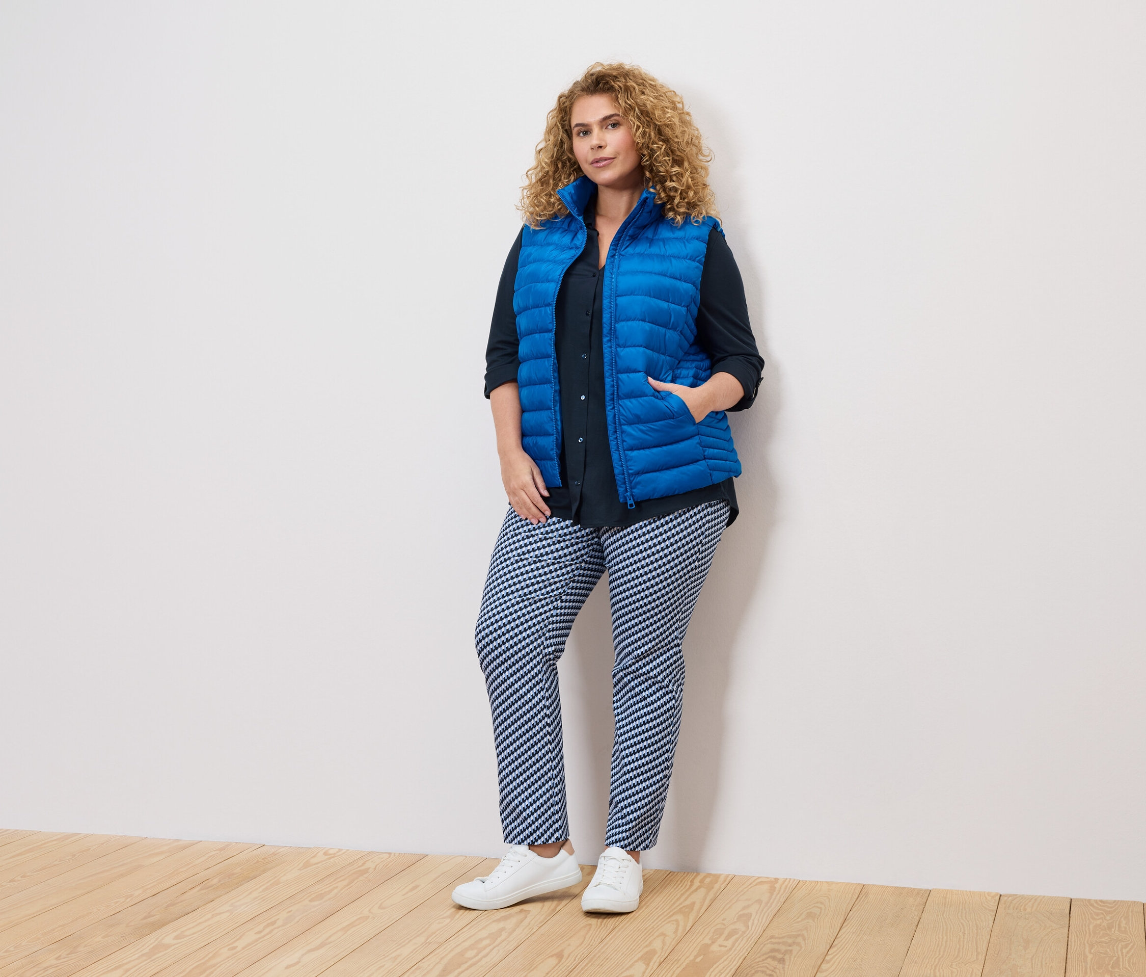 Žena s kudrnatými vlasy v modré vestě, tmavé košili a vzorovaných kalhotách stojí u bílé zdi.