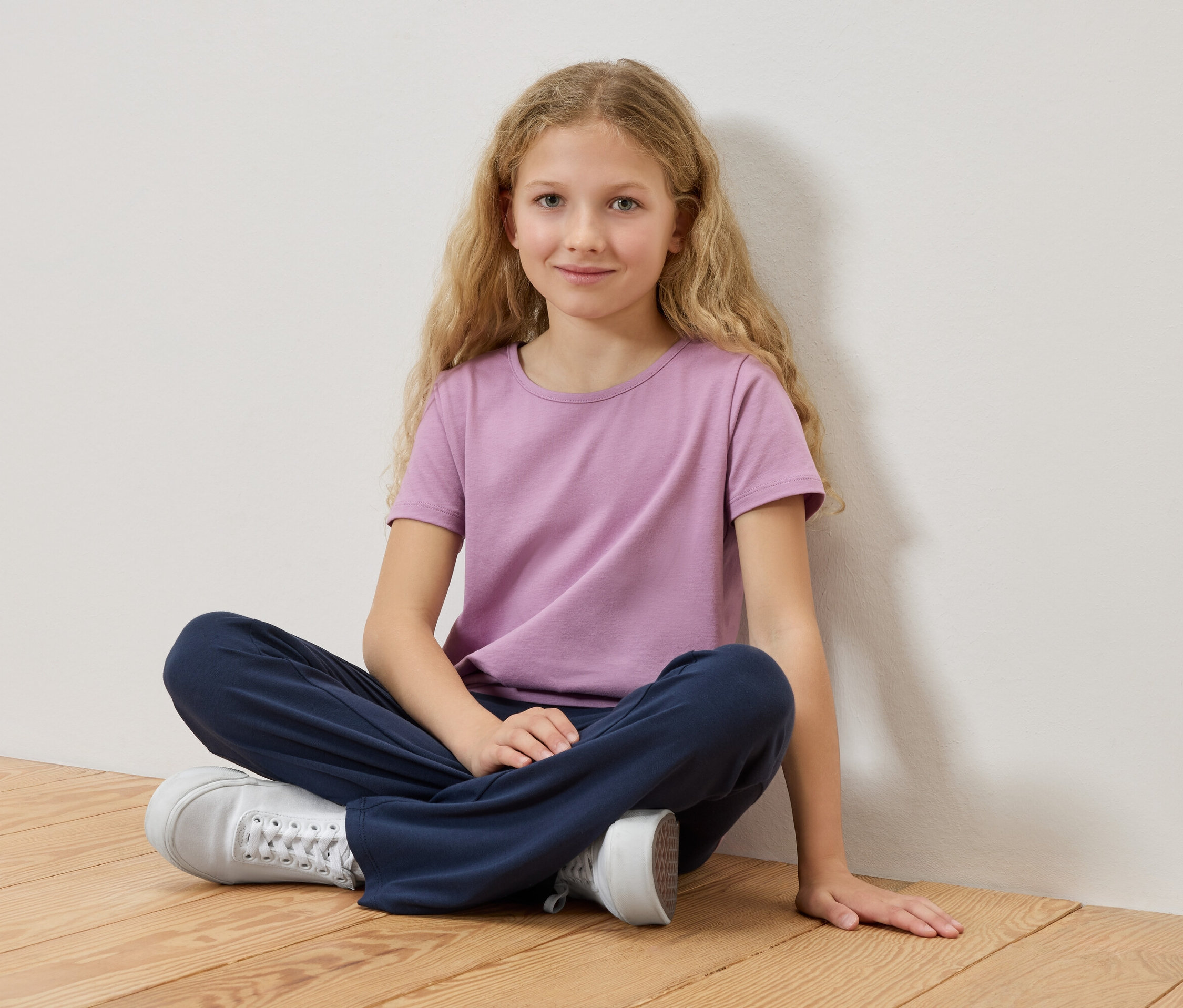 Dívka s blond vlasy sedí se zkříženýma nohama na dřevěné podlaze, má na sobě fialové tričko a modré kalhoty.