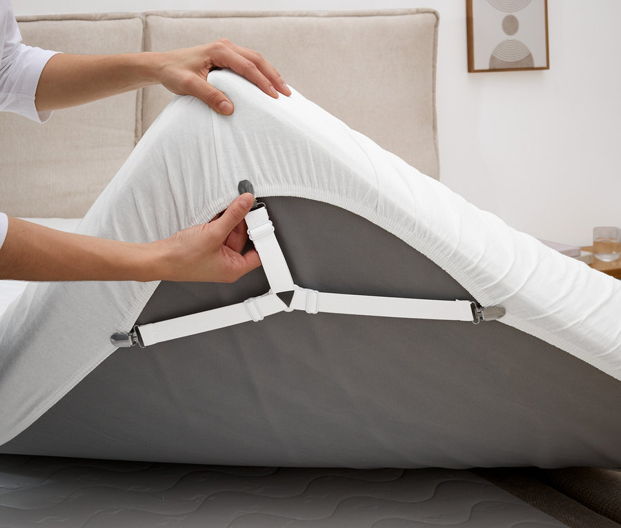 Osoba připevňuje bílý napínák prostěradla na šedé napínací prostěradlo na matraci.