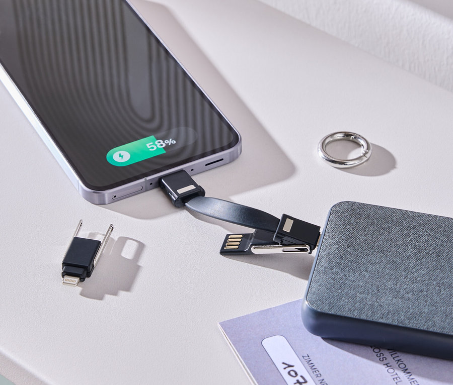Smartphone se nabíjí pomocí kabelu a šedé baterie na bílém stole. Vedle leží prsten a karta od pokoje.