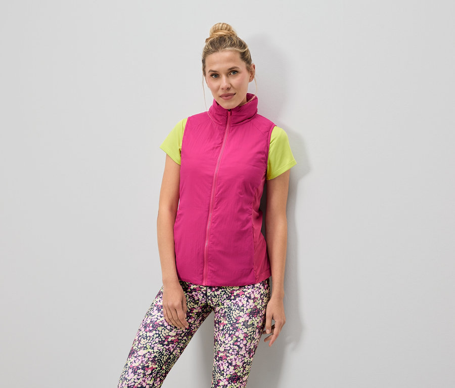 Žena má na sobě růžovou vestu přes světle zelené tričko a květinové legíny.