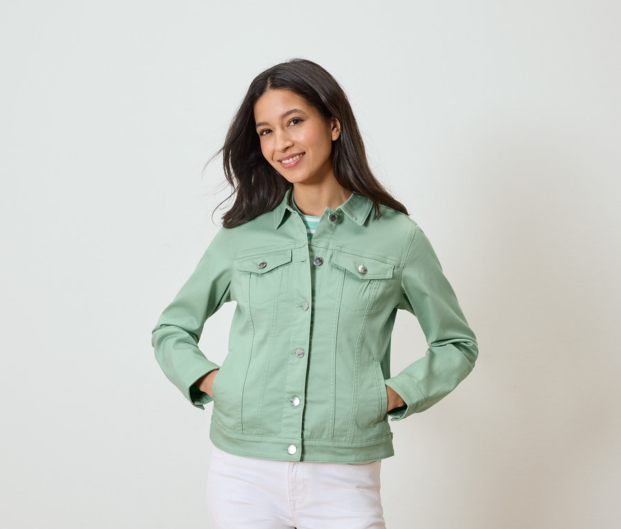 Žena pózuje s rukama v kapsách, oblečená do světle zelené džínové bundy a bílých kalhot.