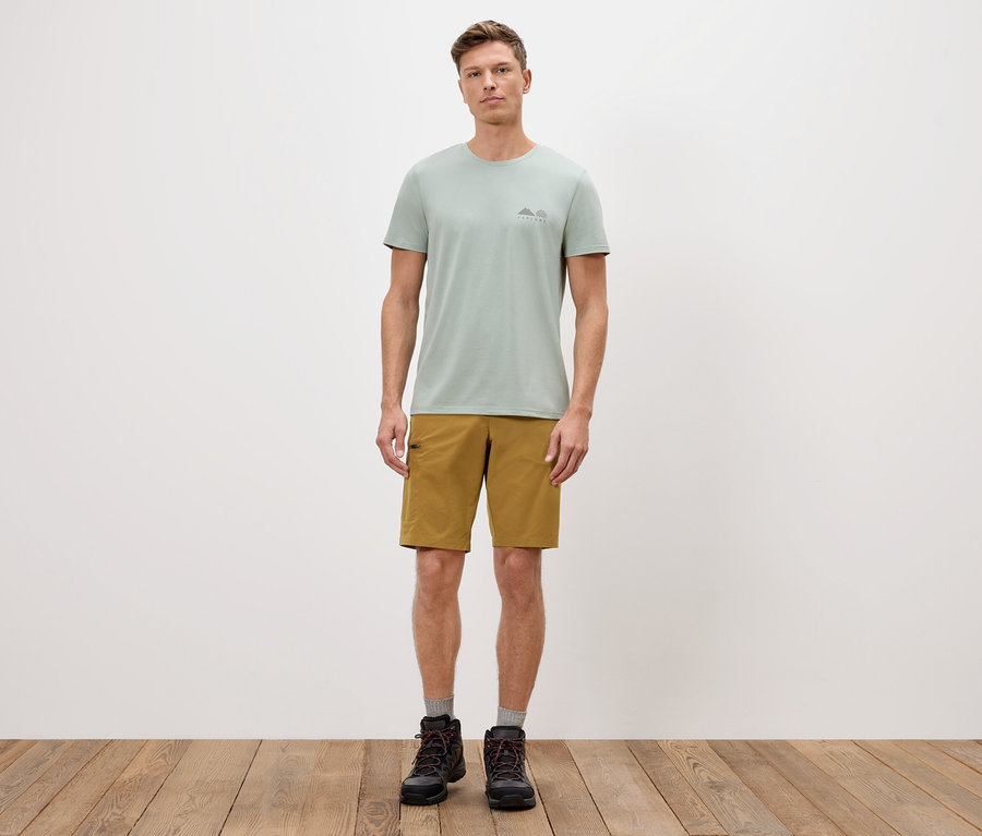 Muž stojí před bílou zdí a má na sobě zelené tričko, šortky a boty.