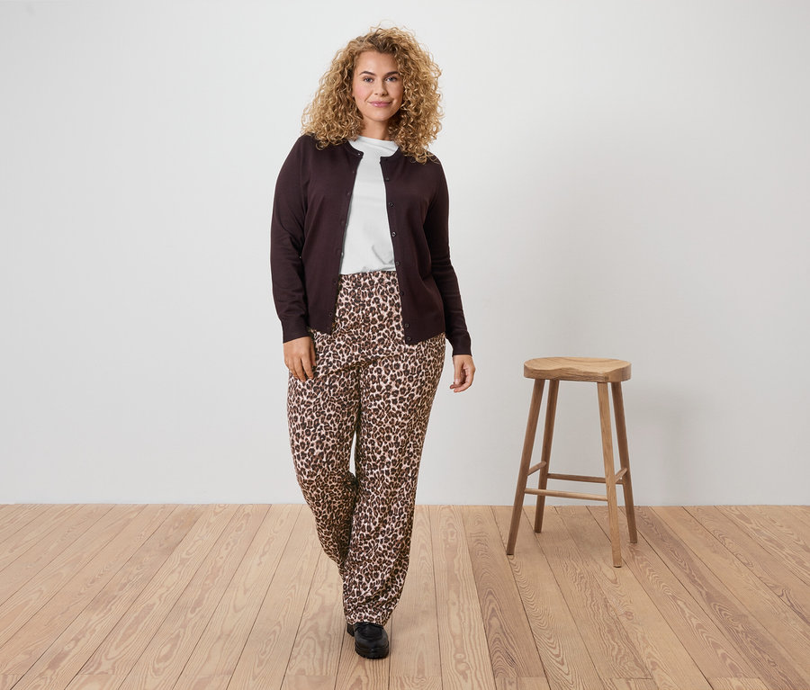 Žena s kudrnatými vlasy má na sobě hnědý kardigan, bílé tričko a kalhoty s leopardím vzorem.