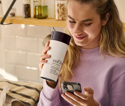 Žena drží v ruce cestovní hrnek na kávu Coffee Dictionary a dívá se na mobilní telefon.