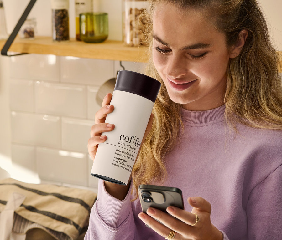 Žena drží v ruce cestovní hrnek na kávu Coffee Dictionary a dívá se na mobilní telefon.