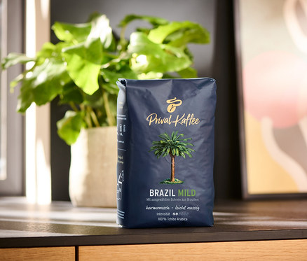 Balení zrnkové kávy Privat Kaffee Brazil Mild 500 g na dřevěné desce. V pozadí je rostlina v květináči a abstraktní obraz.