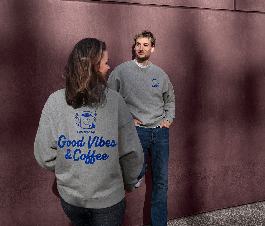 Žena a muž v šedých unisex mikinách Coffee Crew. Na zádech mikiny ženy je modrý nápis "Powered by Good Vibes & Coffee". Muž se dívá na ženu, jeho mikina má modrý nápis "Cheers".
