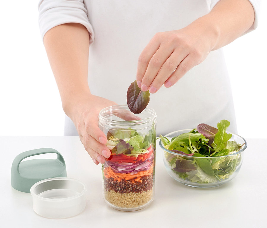 Osoba přidává list salátu do cestovní dózy na oběd, vedle je miska salátu.