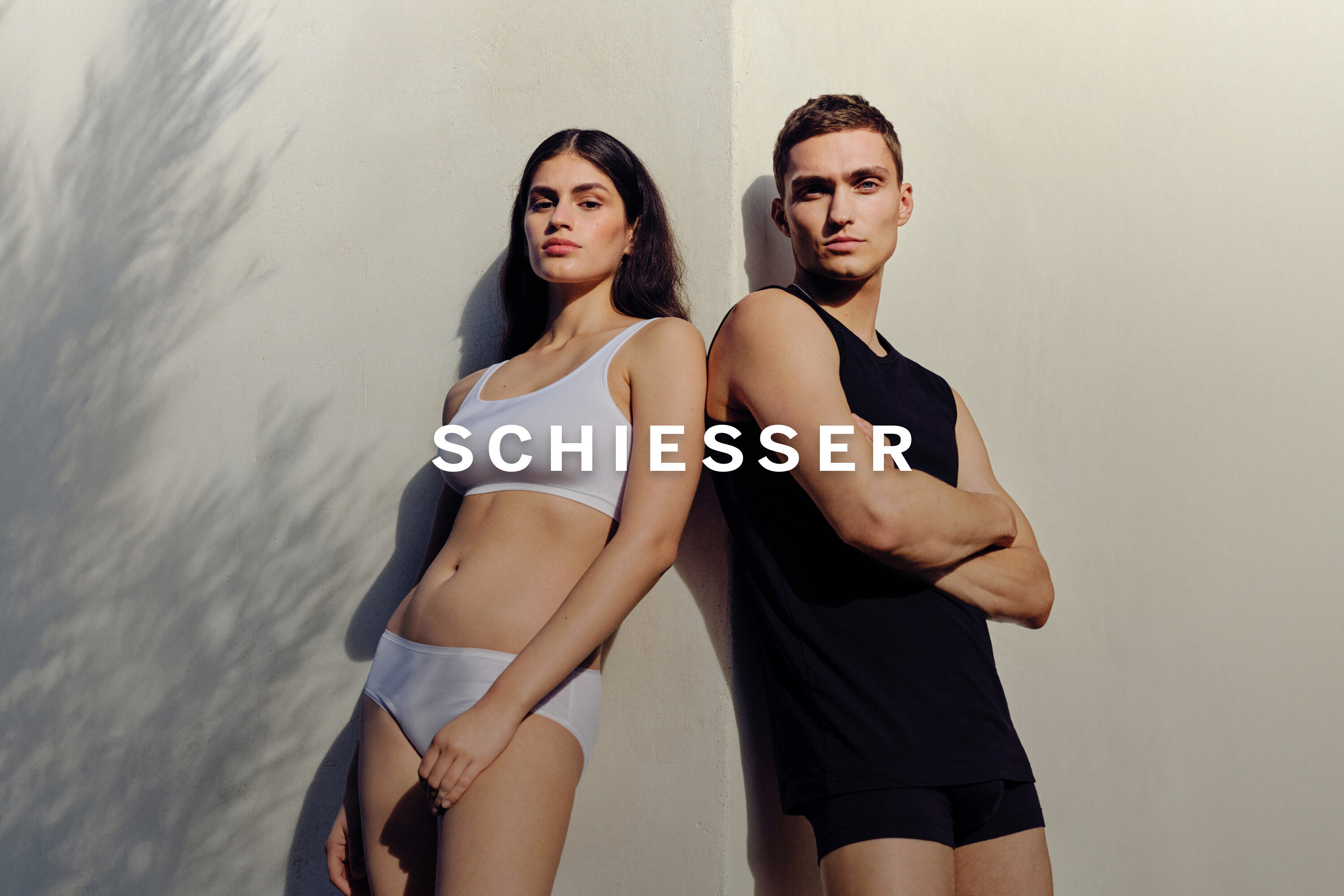 Žena v bílé sportovní podprsence a kalhotkách se opírá o zeď vedle muže v černém tílku a slipách.