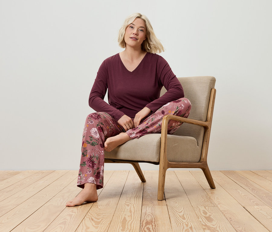 Blondýna sedí v křesle, má na sobě bordó tričko s dlouhým rukávem a pyžamové kalhoty s květinovým vzorem.