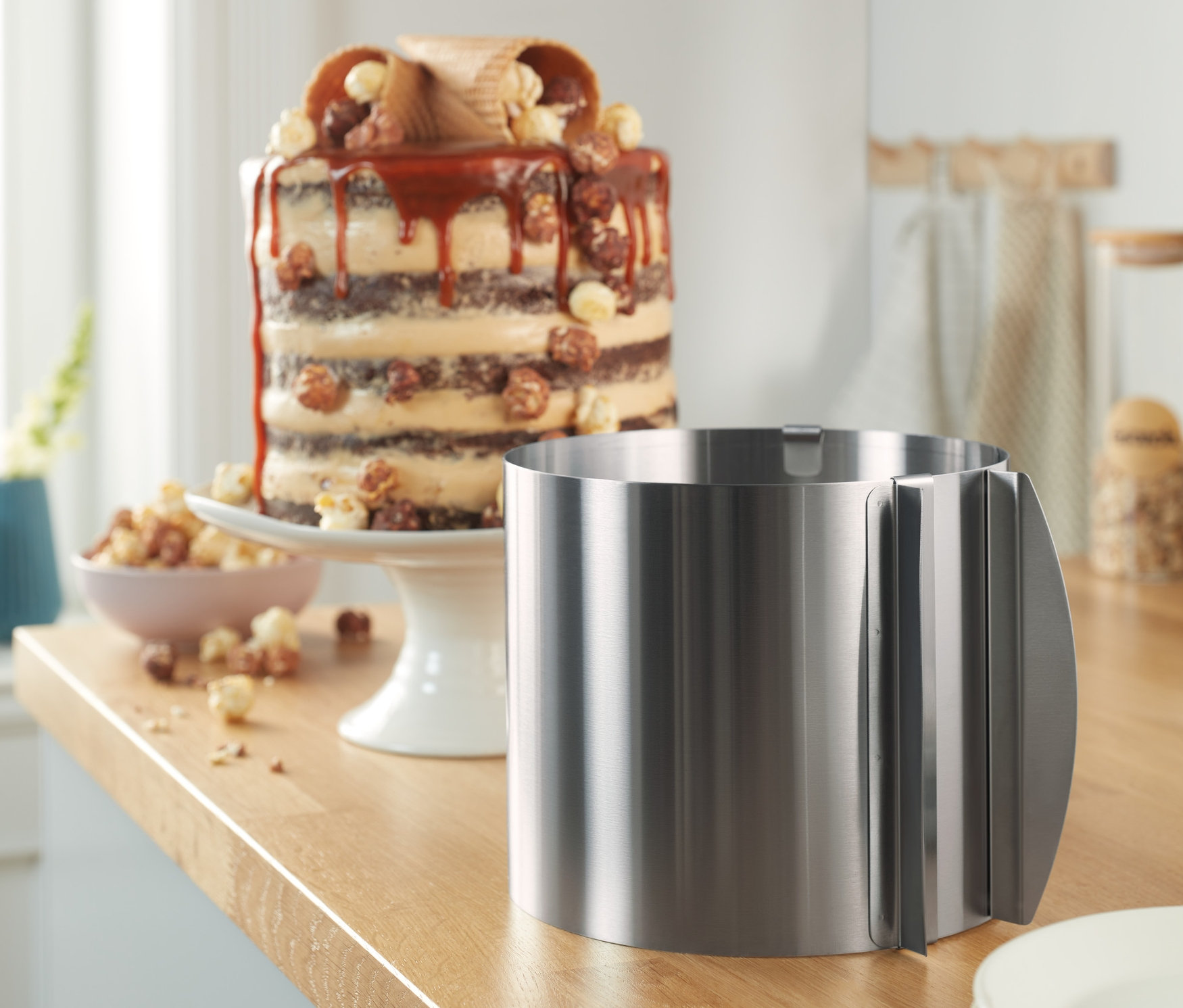 Vysoká forma na dort z ušlechtilé oceli stojí na kuchyňské lince vedle talířů a dortu.