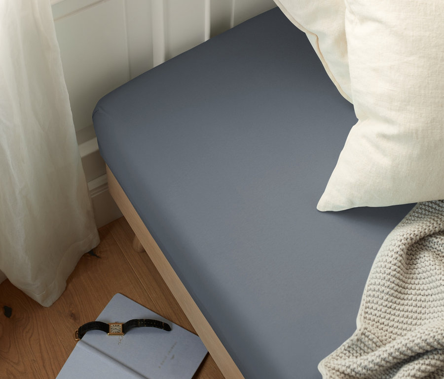 Postel s šedým jersey prostěradlem, dvěma bílými polštáři a pletenou dekou. Na podlaze leží zápisník a hodinky.