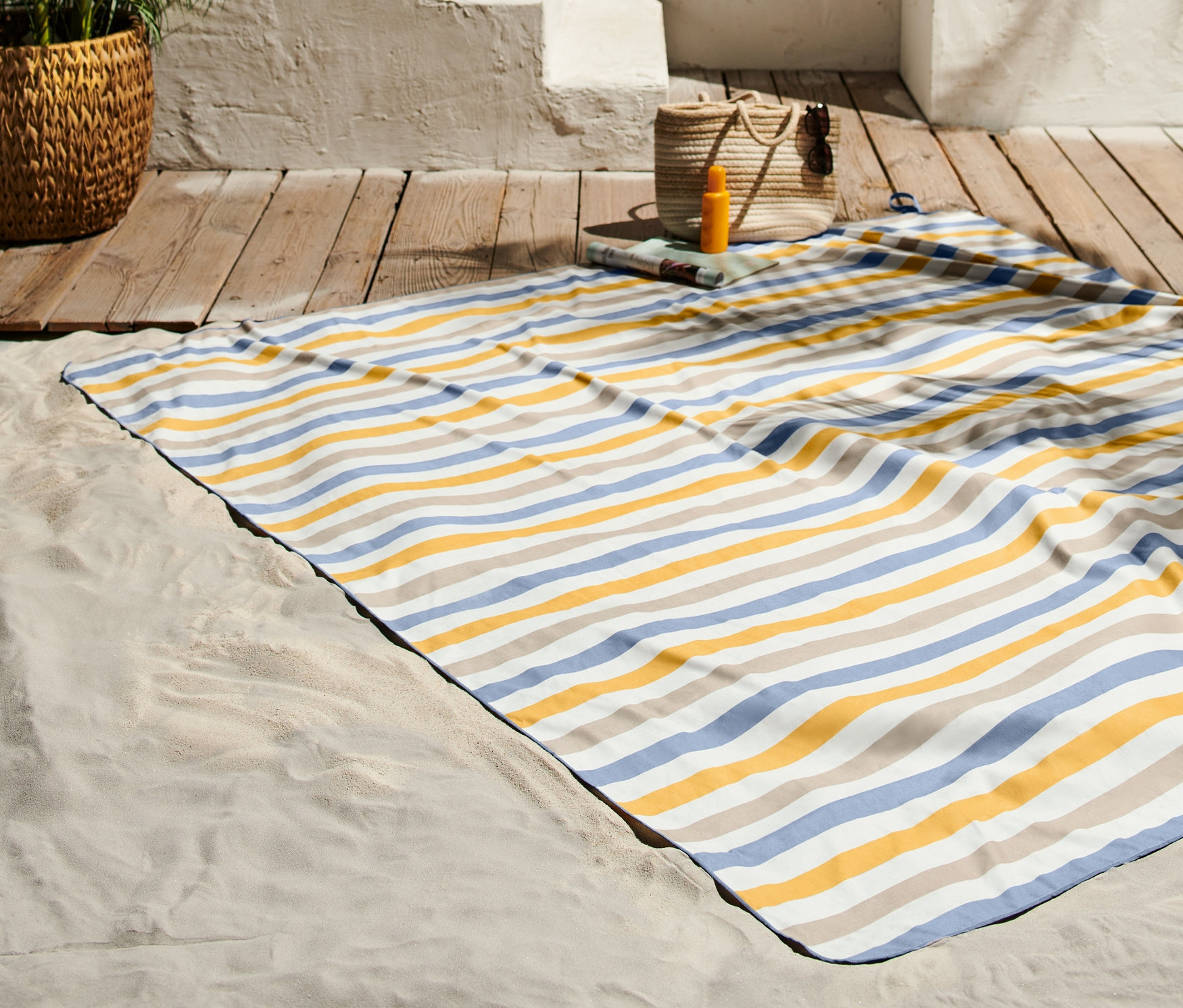 Pruhovaná plážová osuška XL leží na písku vedle dřevěné podlahy.