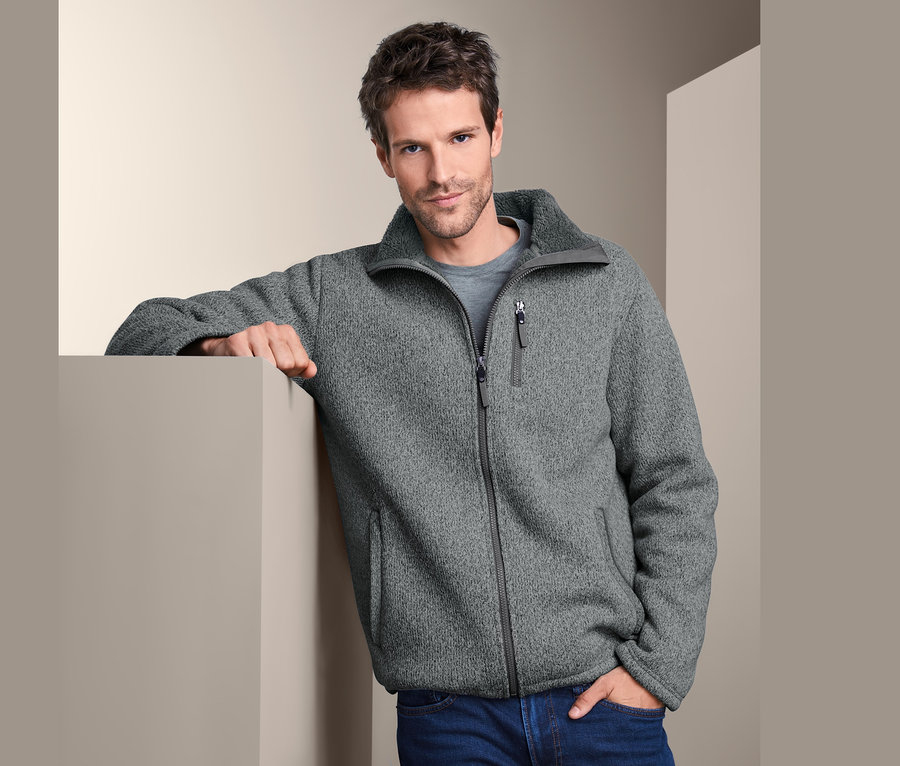 Muž opřený o zeď má na sobě termobundu z pleteného fleecu a džíny.