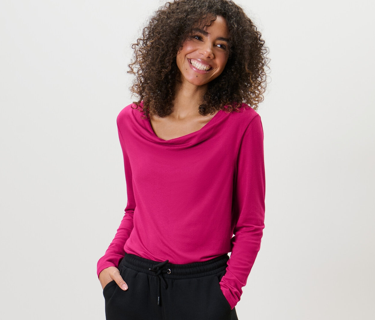 Žena s kudrnatými vlasy nosí halenkové triko s vodopádovým výstřihem a černé kalhoty.