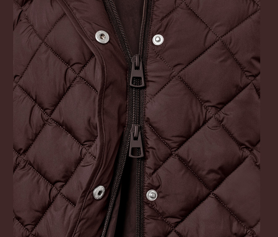 Detailní pohled na dlouhou prošívanou vestu s kapucí, zapnutou na zip a patentky.