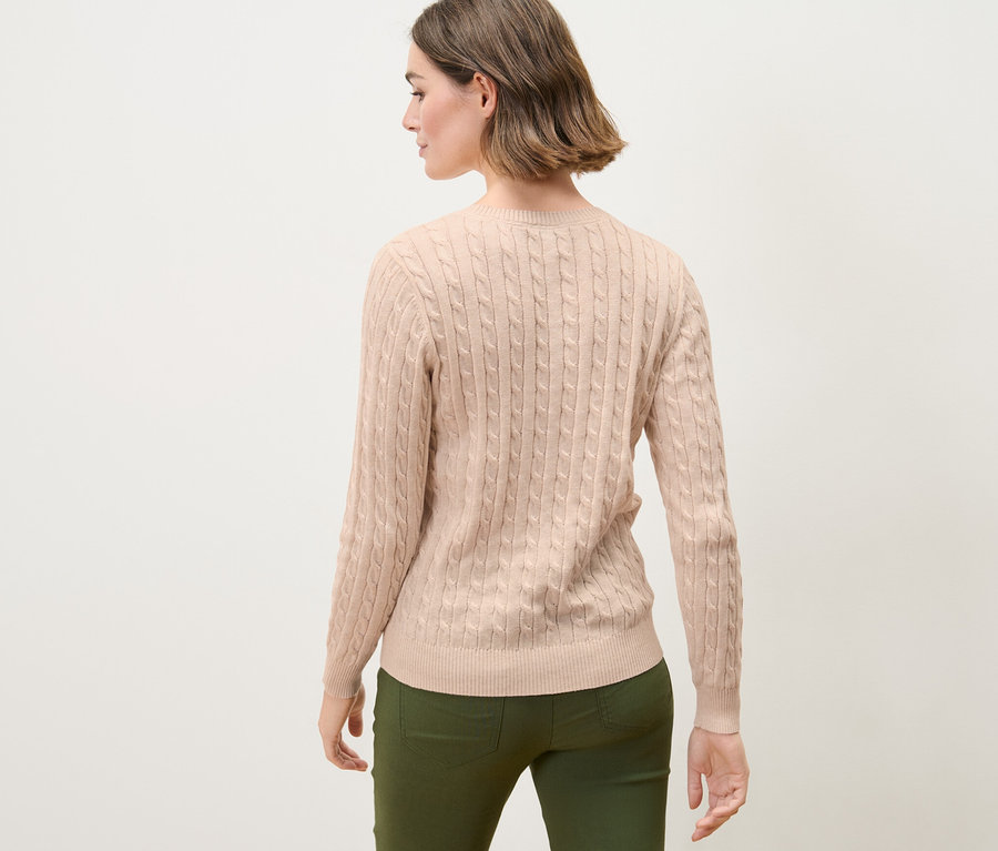 Žena s hnědými vlasy pózuje zády ke kameře a má na sobě krémový svetr s copánkovým vzorem.