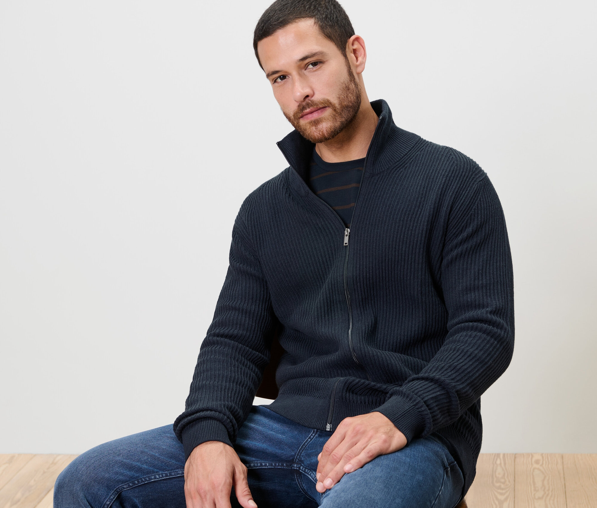 Muž sedí na židli a má na sobě pletený svetr námořnické modré barvy.
