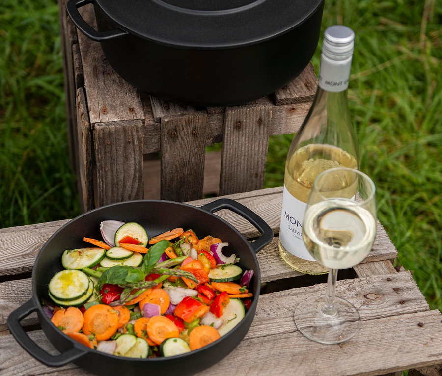 Na dřevěné bedně stojí litinová servírovací pánev COMBEKK Sous-Chef plná zeleniny, vedle ní láhev vína a sklenice vína. V pozadí je další litinový hrnec s poklicí.